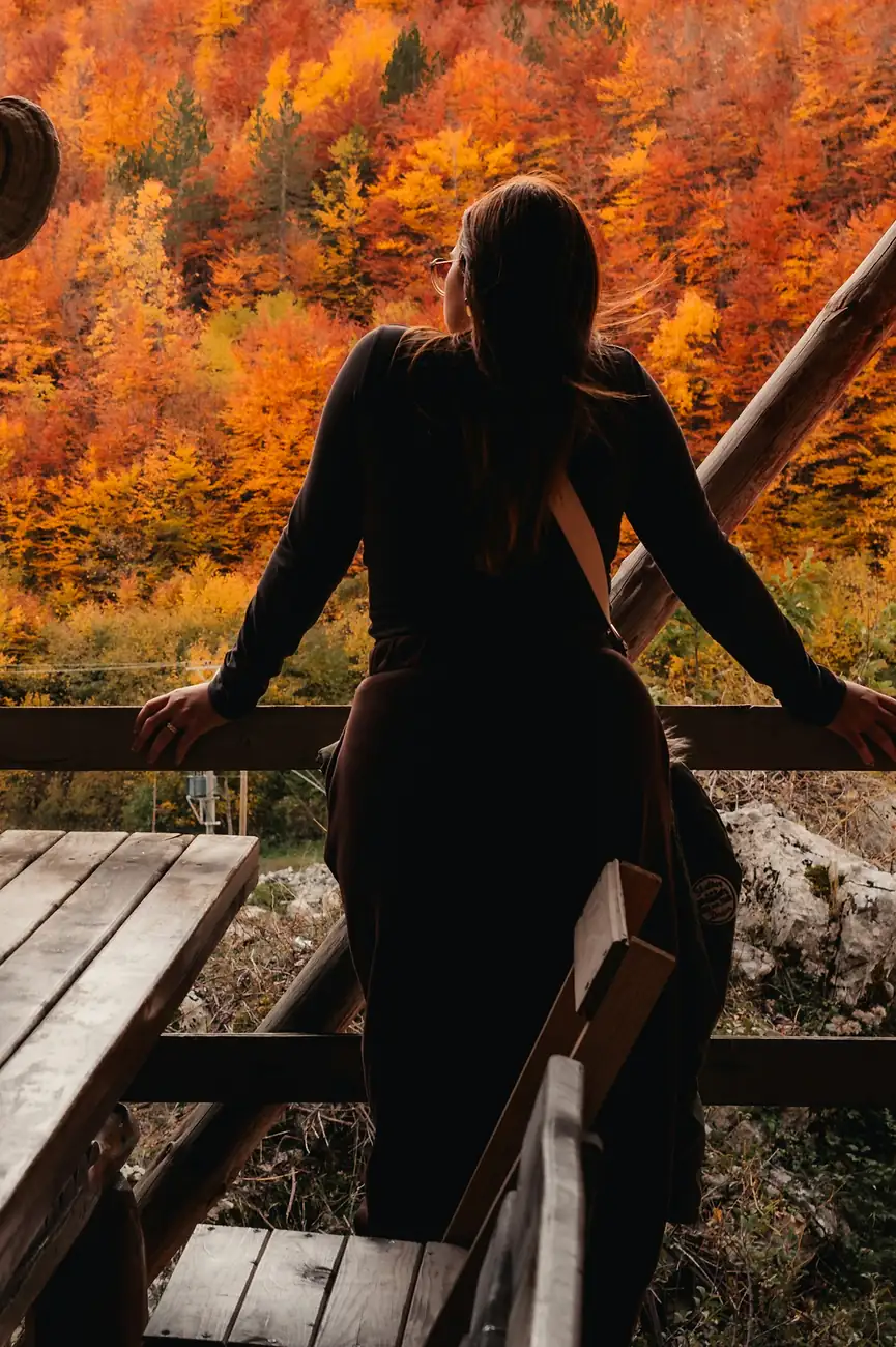 A woman leans on the rail of a wooden terrace, looking out over a hillside of orange autumn trees.
