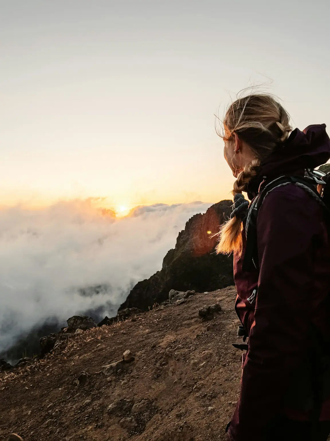 A walker in a dark jacket watches the sun rise over a sea of cloud from a rocky ridge.