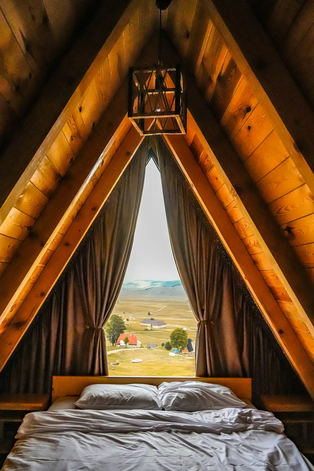 A small A-frame bedroom lined in warm wood, a low double bed with grey linen under a tall window onto the valley.