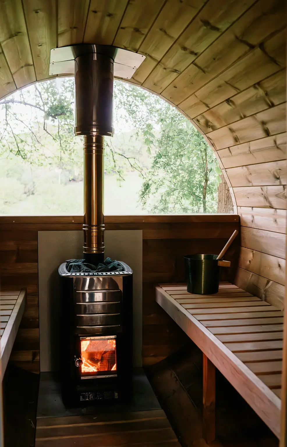 Inside a wooden barrel sauna: a wood-fired stove glowing beneath its chimney, slatted benches and a round window onto birch trees.