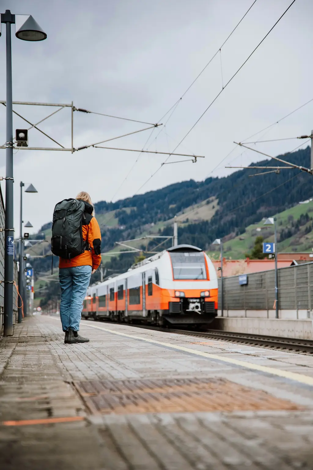 A walker with an orange jacket and a big rucksack waits on a mountain platform as a train pulls in.